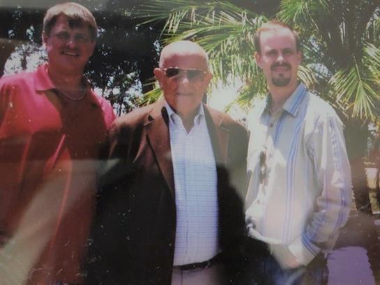 Three friends stand together smiling in a bright, sunny outdoor setting with palm trees.