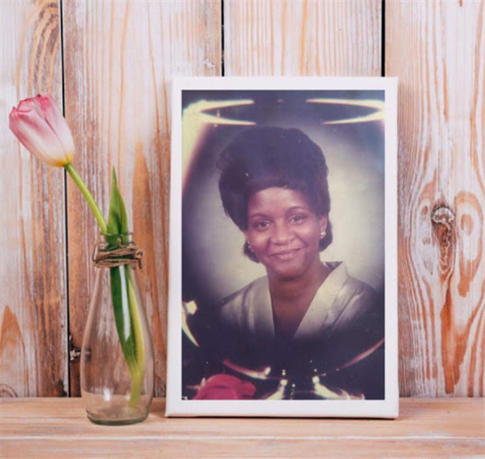 A framed portrait featuring a smiling woman is placed next to a tulip in a glass vase on wood.
