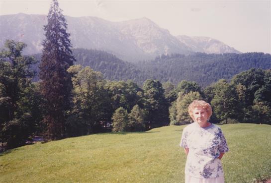 Elderly woman enjoys a tranquil moment in a scenic landscape with mountains and trees around her.