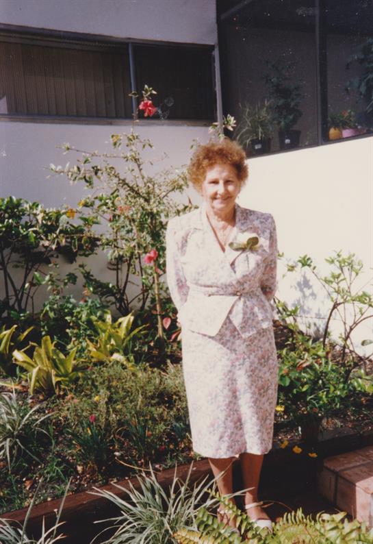 A woman with curly hair stands in a lush garden filled with colorful flowers and greenery.