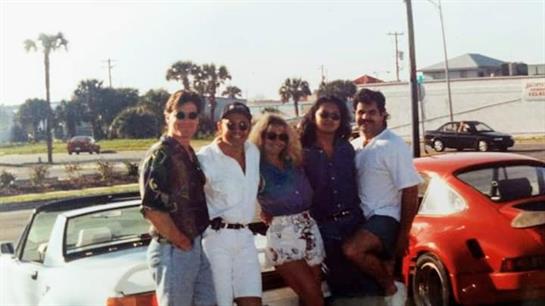 A cheerful group of friends stands together near a classic car, enjoying the warm sunlight.