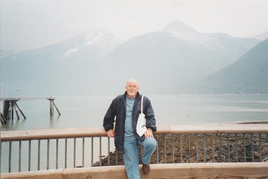 An older gentleman stands by a wooden rail overlooking a calm lake surrounded by mountains.