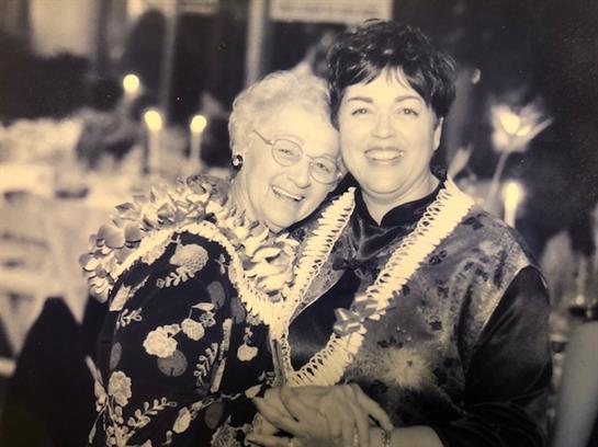 Two women in floral leis and elegant outfits smile while embracing at a festive party.