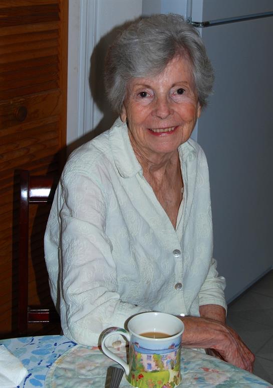 Elderly woman smiles while sitting at a table with a cup in a cozy home environment.