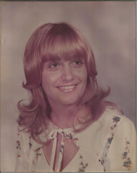 A woman with stylish pink hair smiles at the camera, wearing a floral dress in a studio.