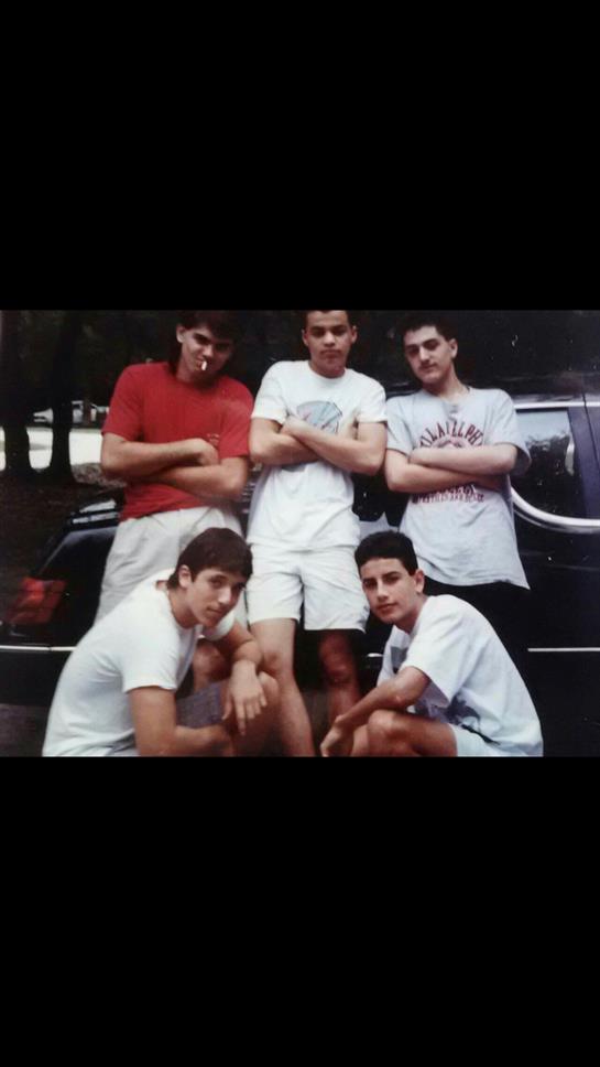 Five young men stand and kneel in relaxed poses near a parked car in a sunny park.