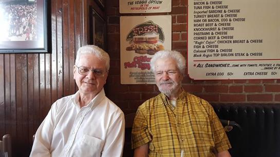 Two older men sit side by side in a cozy restaurant, chatting casually and sharing smiles.