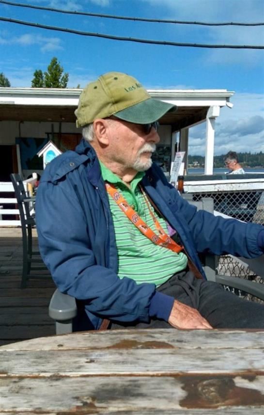 A relaxed elderly man sits in a chair by the water, dressed in casual attire with friends nearby.