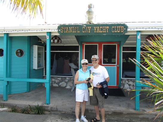 A couple stands smiling outside a colorful yacht club, enjoying their day under clear skies.