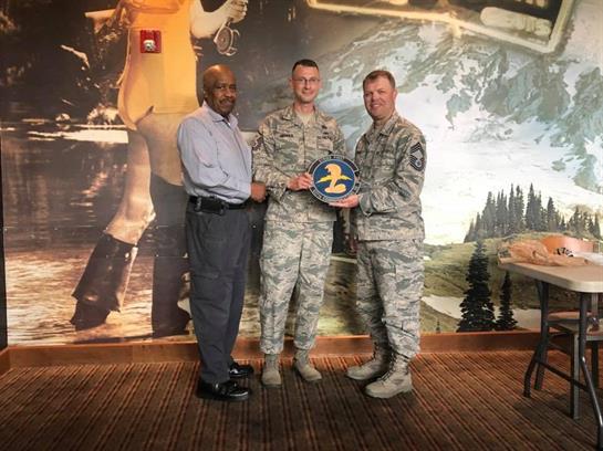 Two service members proudly hold an award while standing with an organizer in a decorated hall.