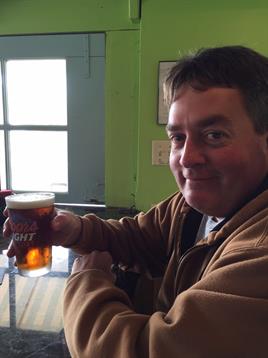 A man smiles while holding a glass of beer at a bar. The atmosphere feels warm and inviting.