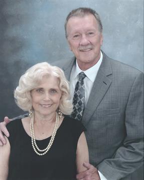 A couple smiles together, both wearing formal outfits, in front of a blue backdrop.
