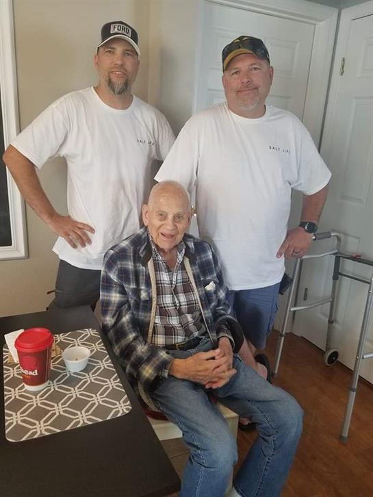 Two men pose confidently behind an elderly man seated, smiling in a warm home environment.