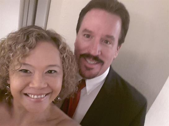 A couple smiles happily while taking a selfie during a formal gathering in a well-lit setting.