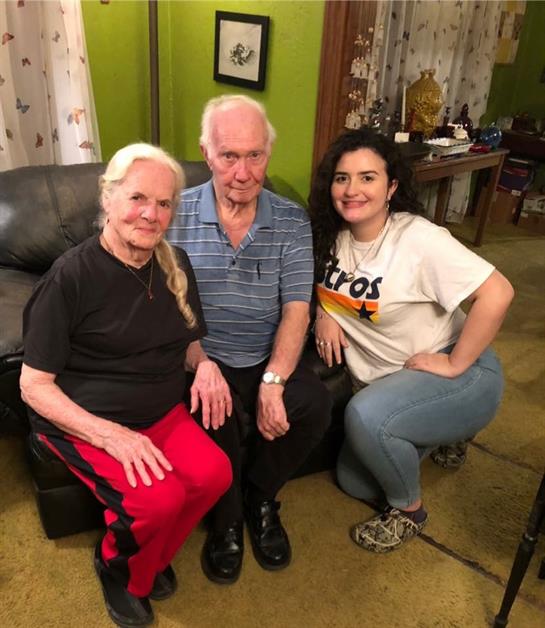 An elderly couple sits on a couch with a smiling young woman kneeling beside them.