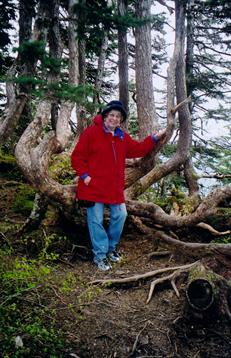 Individual dressed in a red coat poses among trees with exposed roots near a lake.