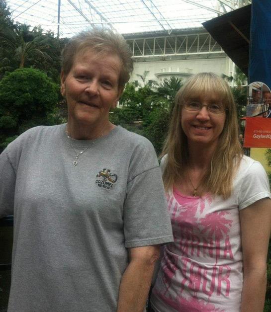 Two women stand close in a lush garden, smiling and enjoying their time outdoors under the sun.