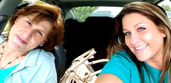 Two friends smile while taking a selfie in a car with a dog sitting behind them.