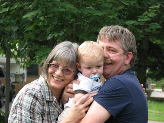 Grandparents embrace their delighted grandchild in a sunny park, sharing a warm family moment.