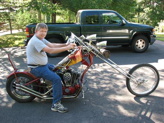 Man sits on an eye-catching chopper bike with extended handlebars in a sunny driveway.