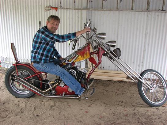 A man in a plaid shirt happily sits on a distinctive motorcycle in a workshop area.