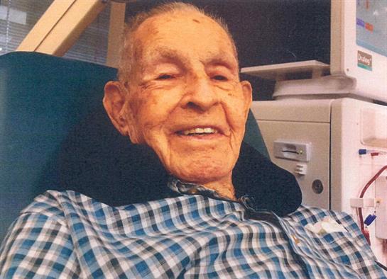 An elderly man with a joyful expression sits comfortably in a chair, wearing a checkered shirt.