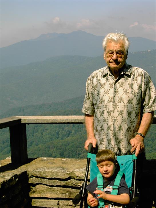 An elderly man stands proudly behind his grandson in a stroller at a scenic viewpoint.