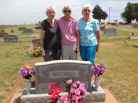 Three women gather around a gravestone, surrounded by flowers, reminiscing and reflecting together.