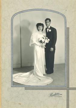 Couple poses elegantly in wedding attire, holding flowers in a neutral indoor location.