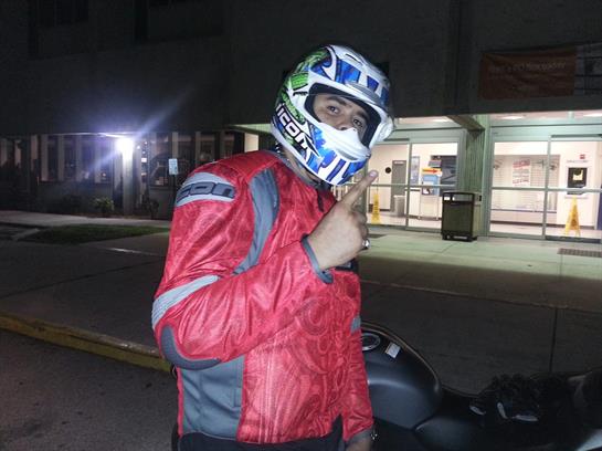 A motorcyclist wearing a helmet and a red jacket gives a thumbs-up gesture in a city at night.