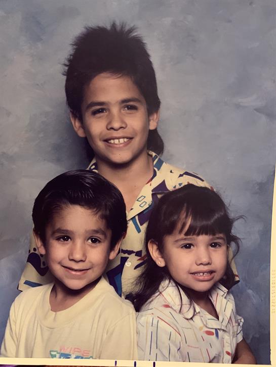 Three children stand together, smiling widely, with playful outfits in a colorful studio.