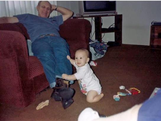 A baby is smiling and reaching for a man's leg while toys are scattered around the floor.
