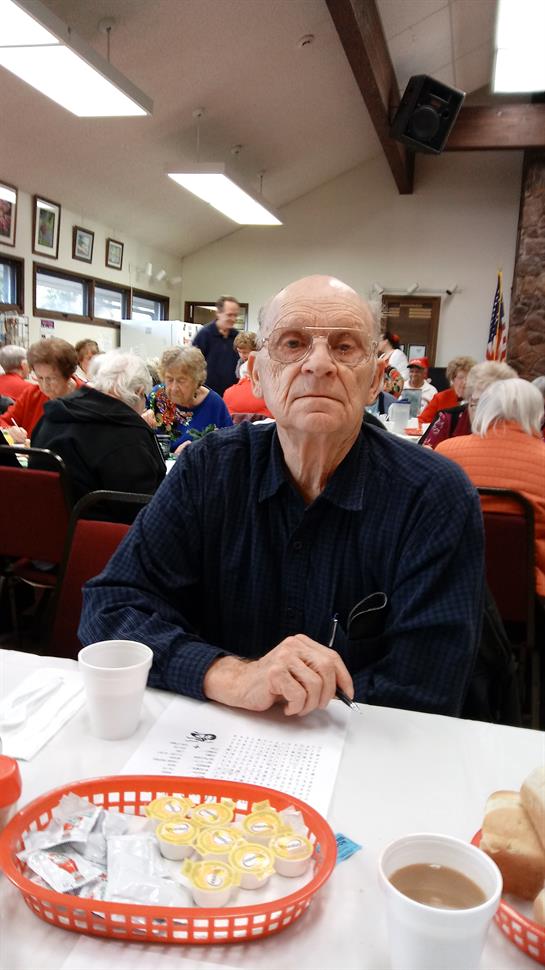 An older gentleman sits at a table in a busy community hall during a social event.