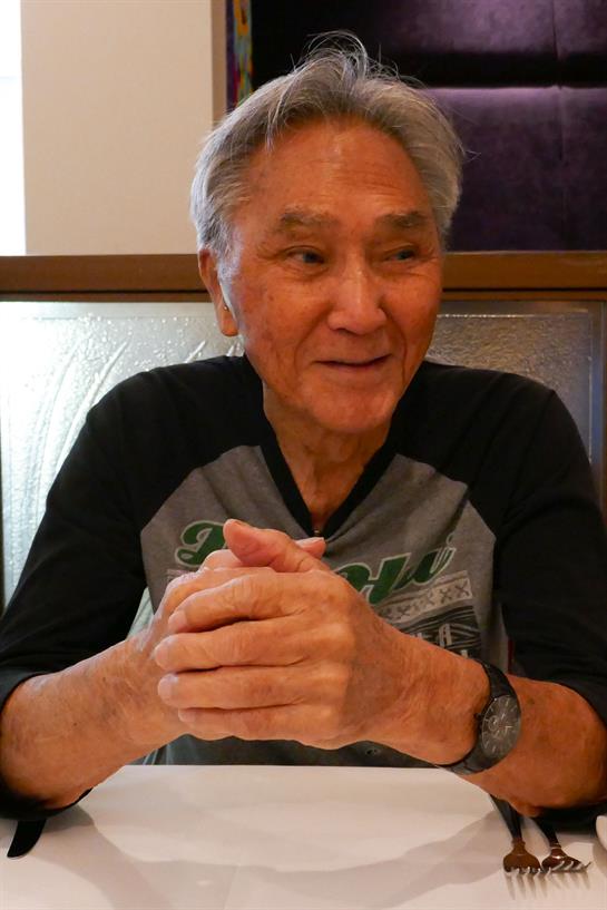 An older man smiles and chats with others at a restaurant table.