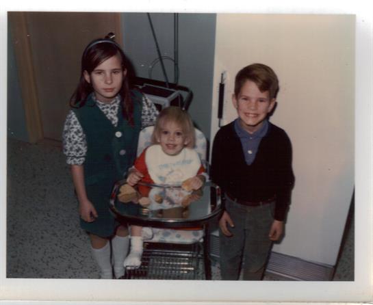 Three children stand close together, smiling in a cozy indoor space with a stroller.