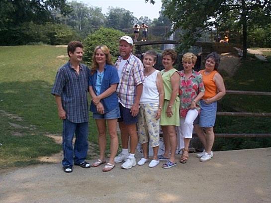 A group of seven people stands together in a park, smiling and enjoying their time outdoors.