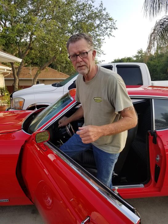 A man is getting out of a classic red car parked in a driveway surrounded by trees.