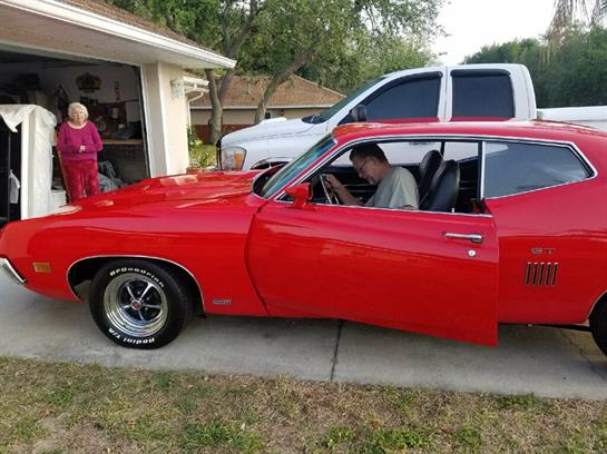 A vintage red muscle car is parked near a garage while a child observes and an adult sits inside.