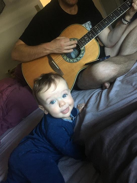A baby smiles while sitting on a bed as someone plays guitar nearby in a cozy room.