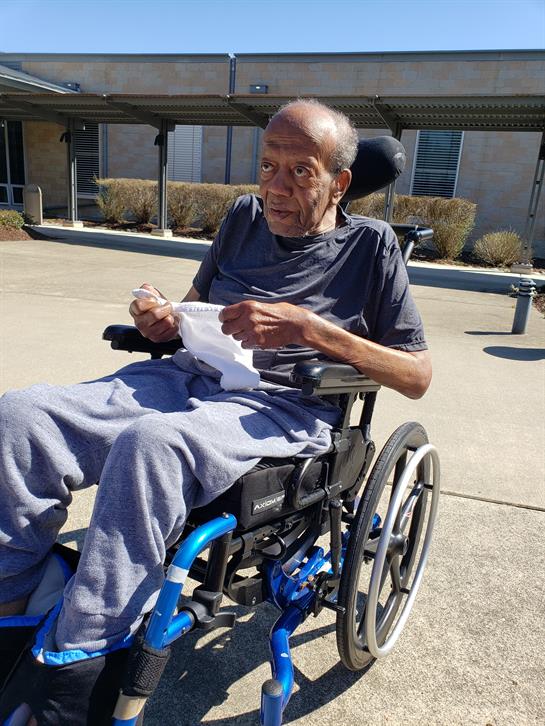 An elderly man sits in a wheelchair, relaxed outdoors at a care facility, enjoying a snack.