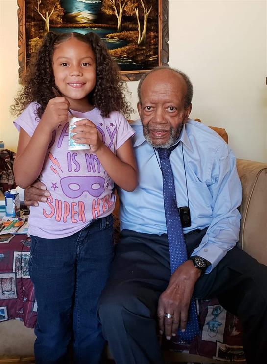 Young girl in casual clothes smiles while holding a snack beside an older man in a blue shirt.