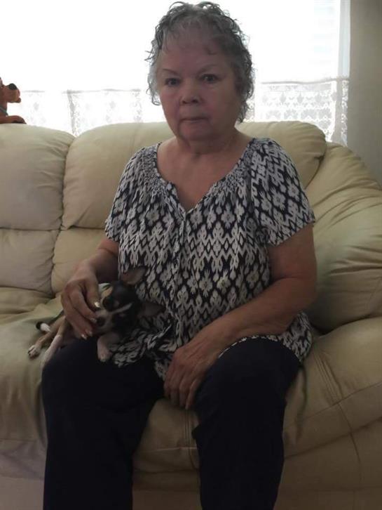 An elderly woman holding a small dog sits thoughtfully in a light-colored living room.