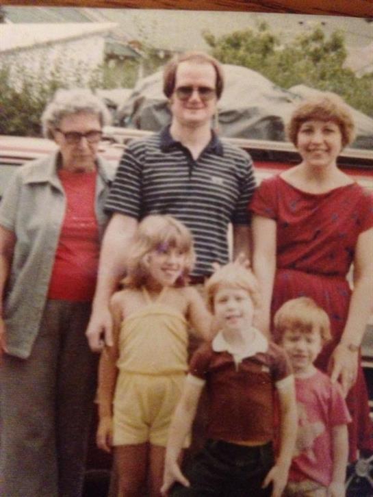 A family poses together outside with a vintage vehicle in the background, enjoying a sunny day.