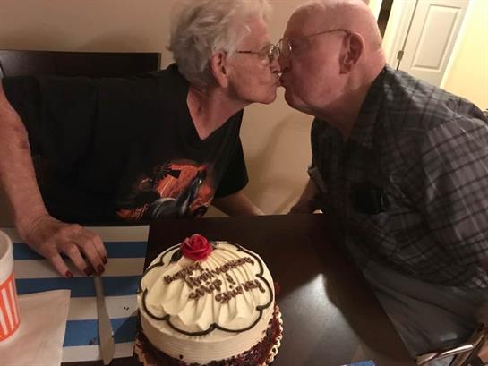 An elderly couple kisses sweetly beside a beautifully decorated cake at their home party.