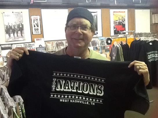 A man in a vintage record store holds a black shirt with The Nations logo visible.