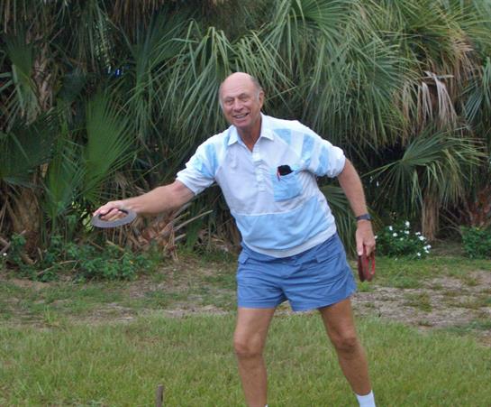 A cheerful elderly man dressed in casual attire throws a frisbee in a vibrant garden.