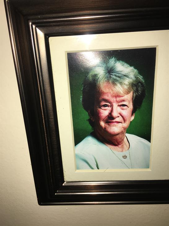 An older woman with gray hair smiles softly against a green background in a framed portrait.