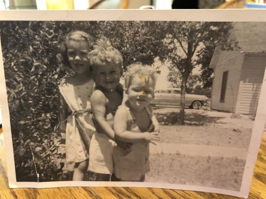 Three young children pose joyfully outdoors in a garden beside a house during the summer.