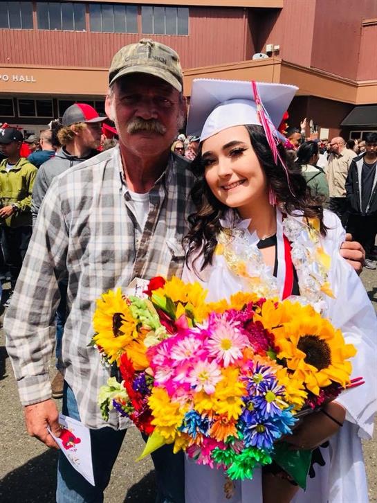 A father and daughter joyfully celebrate her graduation among blooming flowers.
