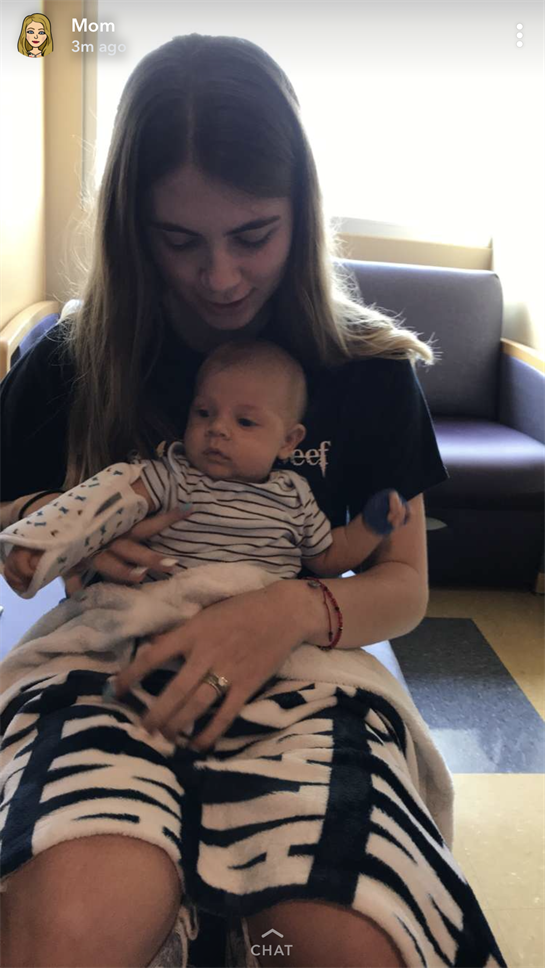 A young woman gently holds a baby in a medical environment while sitting on a chair.